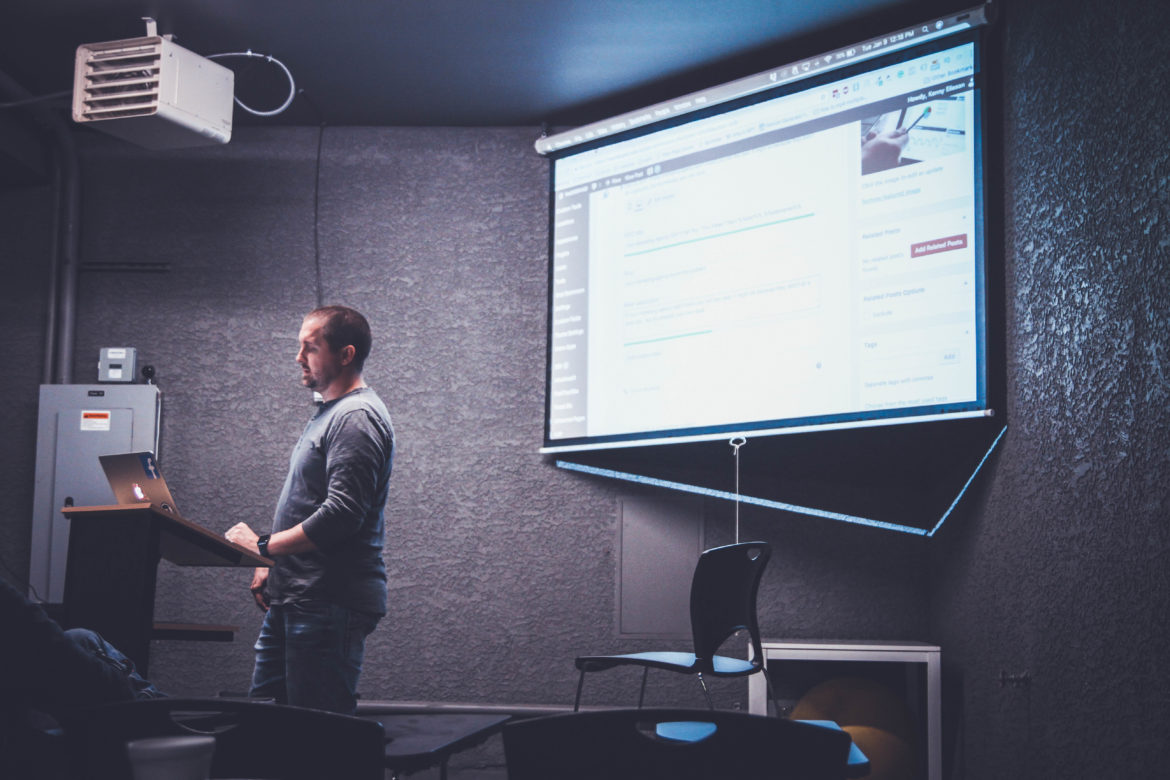 A man using a classroom projector with the image on a screen behind him